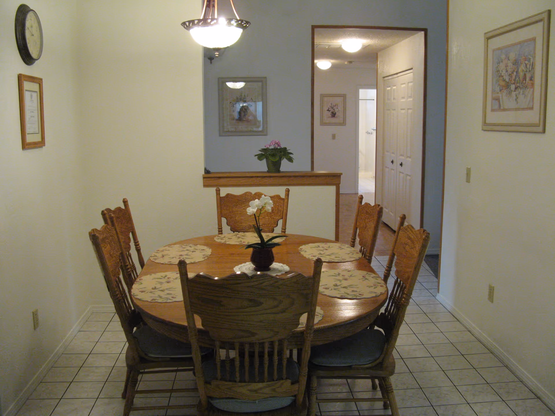 A dining room with a wooden oval table surrounded by six wooden chairs with cushions. The table has six floral placemats and a small potted orchid in the center. The room has tiled flooring, light-colored walls, a hanging ceiling light, a wall clock, and framed pictures. There is an open doorway leading to a hallway with more framed pictures and a glimpse of a bathroom.