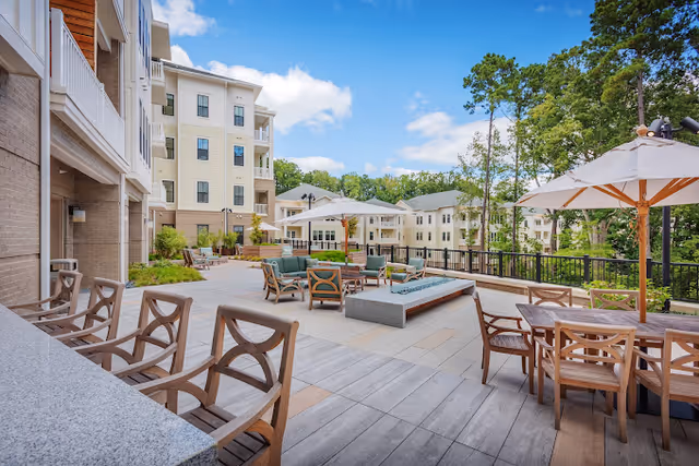 Outdoor patio area at a senior living facility with wooden chairs and tables, large umbrellas providing shade, cushioned seating around a rectangular fire pit, and multi-story residential buildings in the background under a partly cloudy sky.