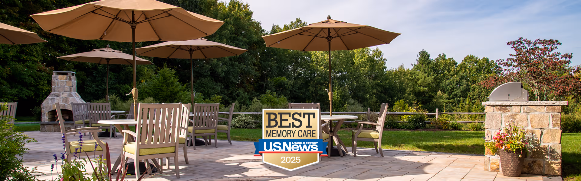 Outdoor patio area with several tables and chairs under large beige umbrellas, surrounded by greenery and trees. There is a stone fireplace on the left and a stone grill on the right with a flower pot. A sign in the center reads 'Best Memory Care U.S. News 2025'.