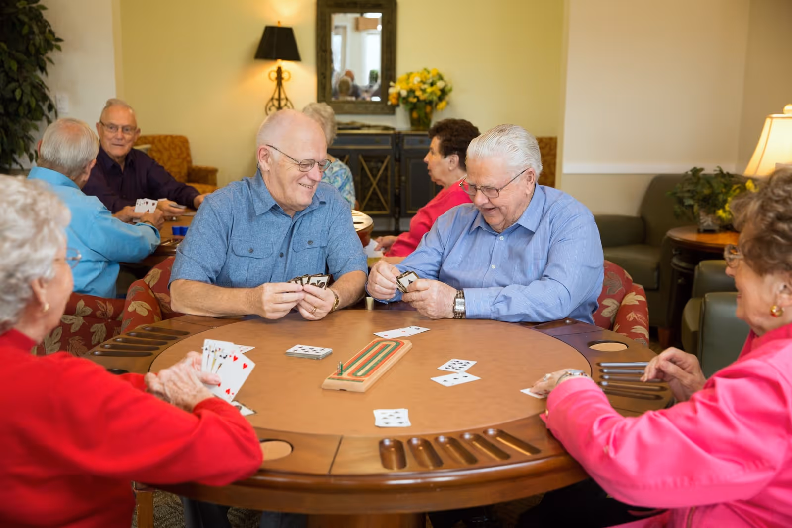 A group of elderly people sitting around a round wooden table playing cards in a cozy room with warm lighting, plants, and a lamp in the background.