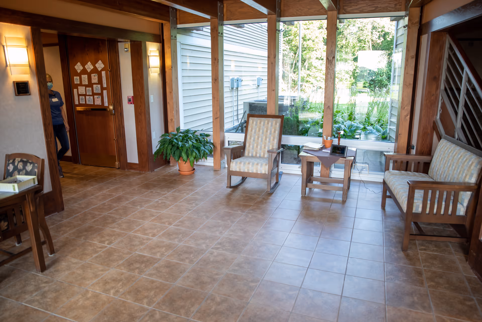 A bright seating area in a senior living facility with tiled floors, wooden framed chairs with patterned cushions, a small wooden table with books and a plant, large windows showing greenery outside, and a person wearing a mask partially visible near a wooden door.
