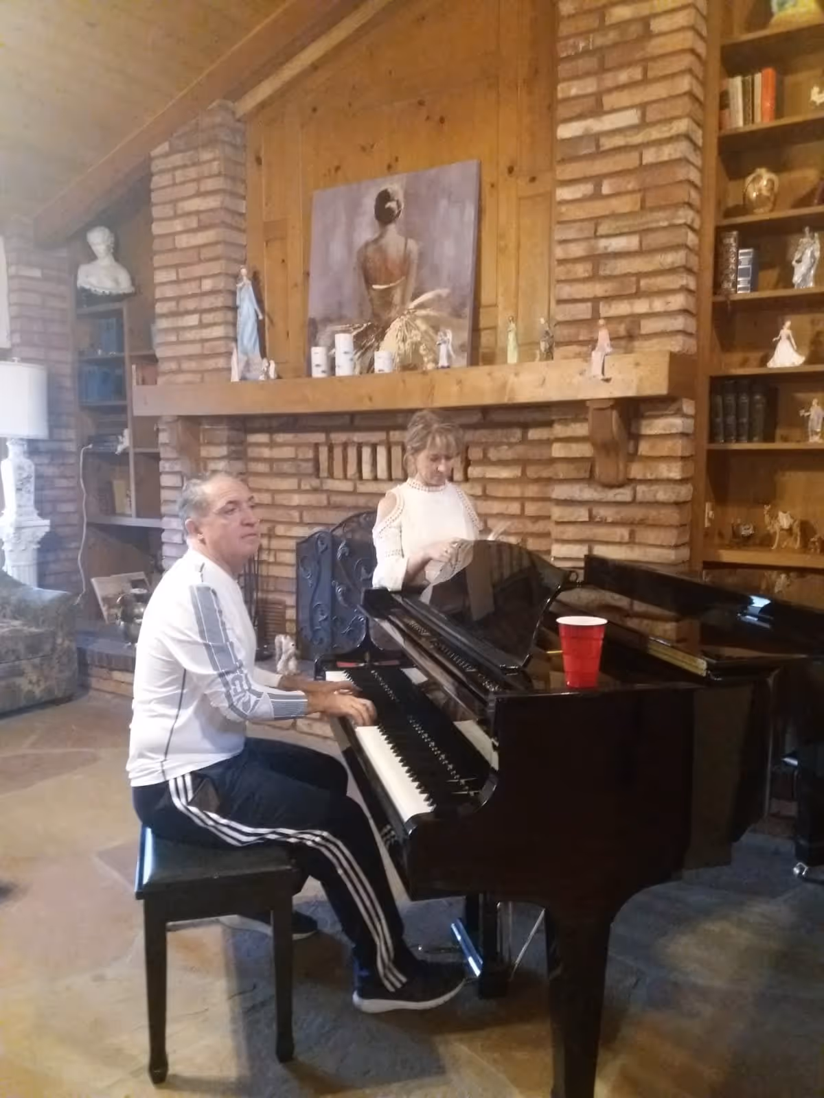 A man playing a black grand piano in a cozy room with brick walls and wooden shelves. A woman stands behind the piano looking at sheet music. The room features a fireplace with a wooden mantel decorated with small figurines and a painting of a woman with her back turned. There is a red plastic cup on the piano and a floral armchair to the left.