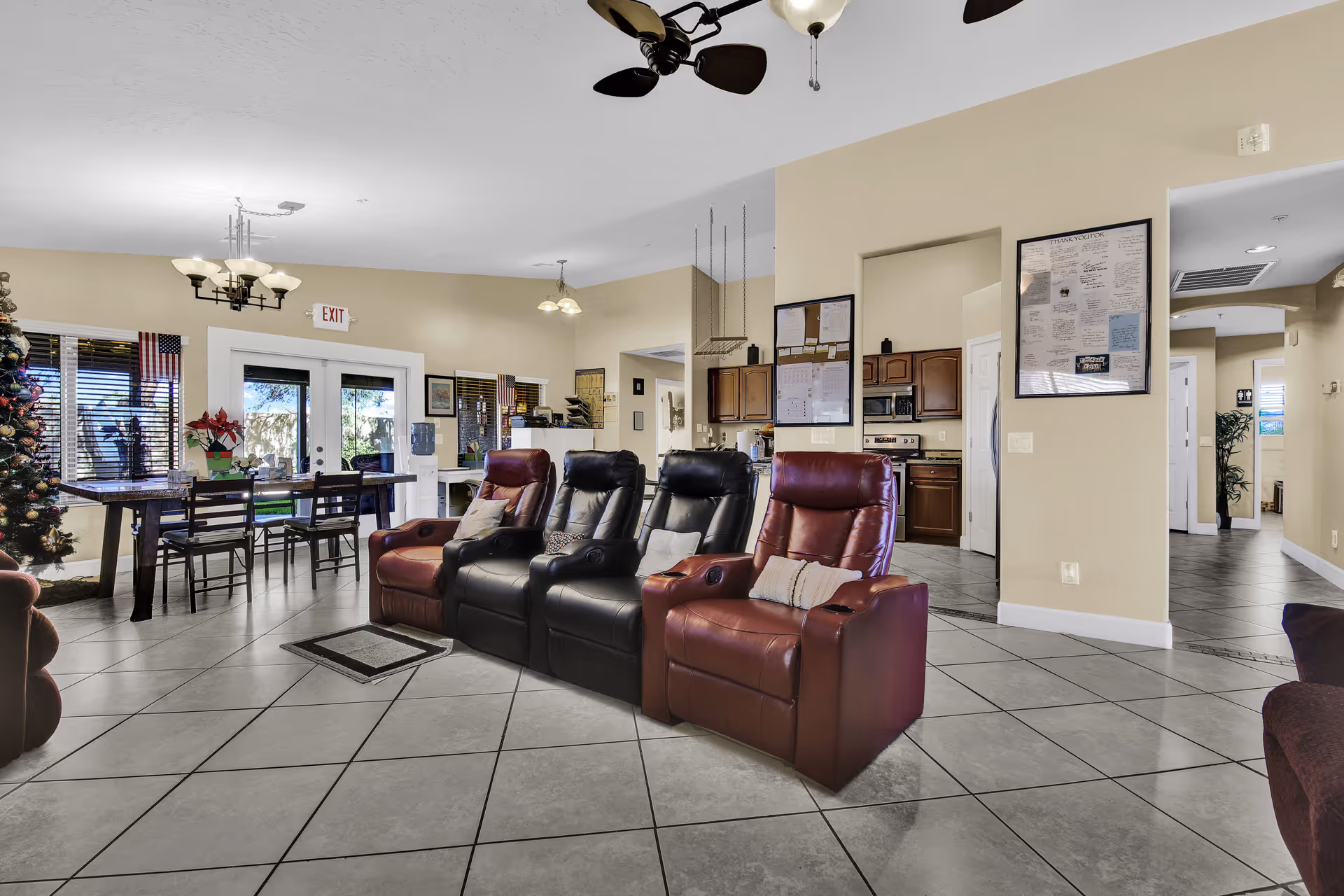 A spacious living area in a senior living facility featuring four recliner chairs in the center, two in dark brown and two in maroon. Behind the chairs is a dining table with chairs near large windows and glass doors leading outside. The room has beige walls, tiled floors, ceiling fans, and light fixtures. A decorated Christmas tree is visible on the left side, and a kitchen area with wooden cabinets and appliances is seen in the background.