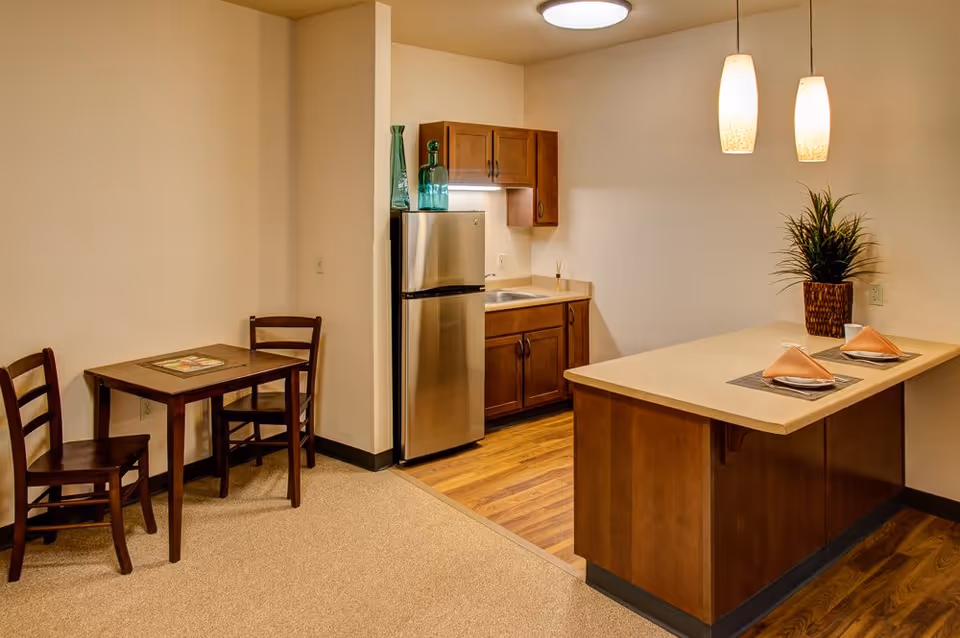 A small kitchen and dining area in an assisted living facility. The kitchen features wooden cabinets, a stainless steel refrigerator, a sink, and a countertop with two place settings and a potted plant. Adjacent to the kitchen is a small wooden dining table with two chairs. The room has warm lighting from ceiling fixtures and pendant lights above the counter.