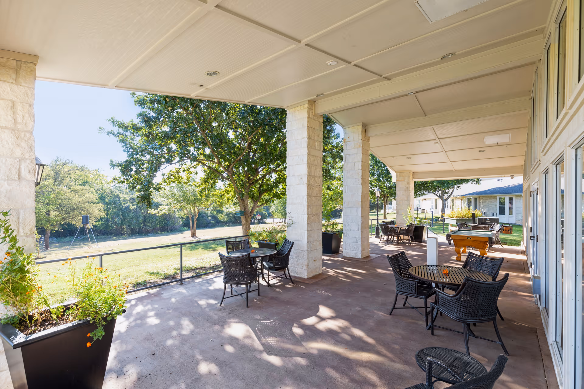 Covered outdoor patio area with multiple round tables and black wicker chairs. Large stone pillars support the roof, and there are potted plants along the edge. Beyond the patio is a grassy area with trees and a clear sky.