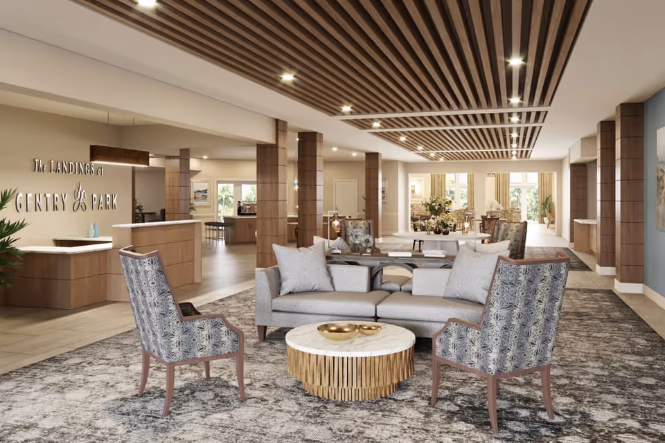 A spacious and modern senior living facility lobby with a seating area featuring a gray sofa, two patterned armchairs, and a round coffee table. The ceiling has wooden slats with recessed lighting. The reception desk is visible on the left with a sign that reads 'The Landings at Gentry Park'. The background shows more seating and large windows letting in natural light.