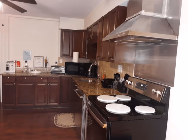 A kitchen with dark wooden cabinets, granite countertops, and stainless steel appliances including a stove with four burners and a large range hood. There is a microwave, toaster oven, coffee maker, and various kitchen utensils on the counter. The floor is wooden with a small rug near the sink area.
