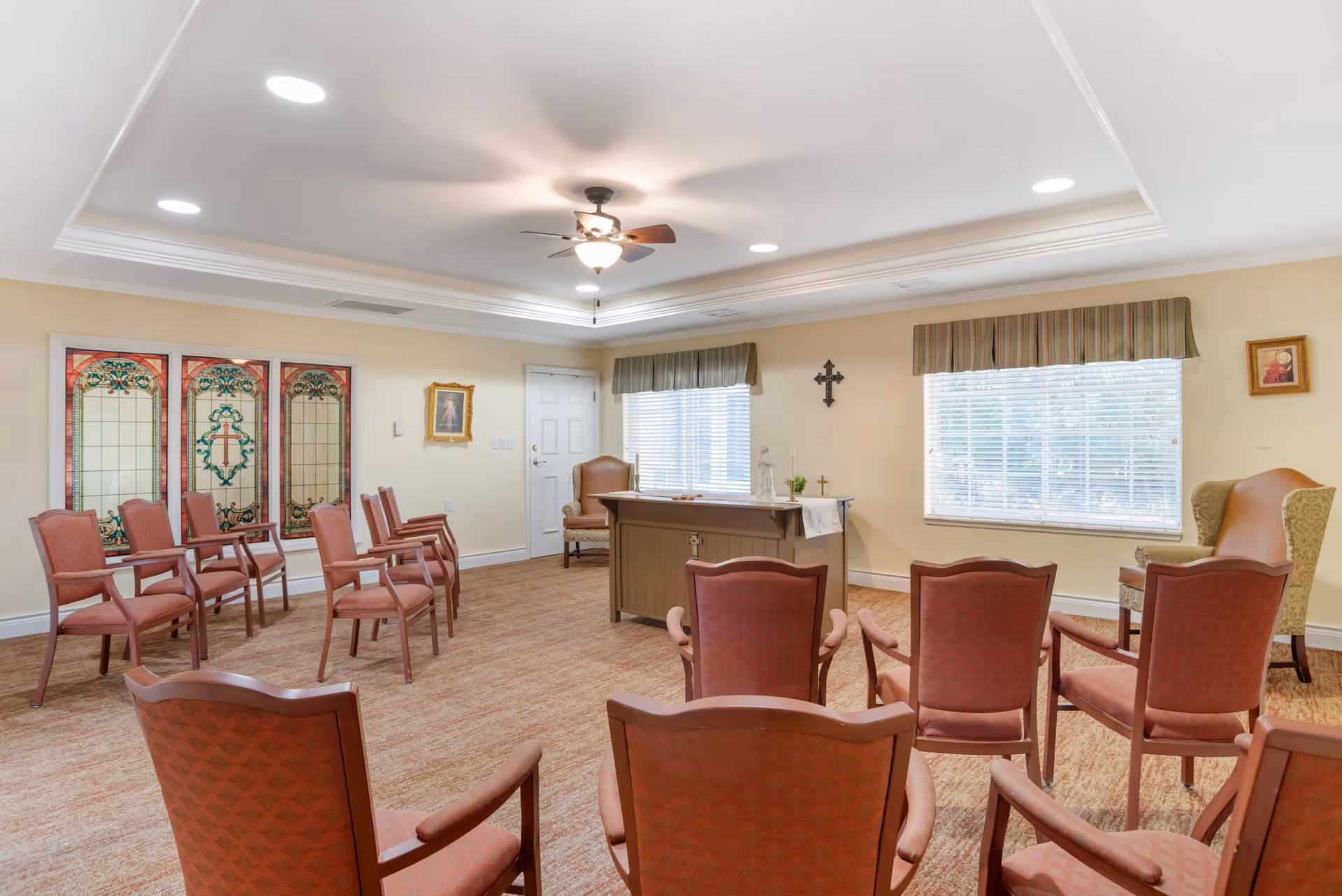 A small chapel or prayer room with rows of red cushioned chairs facing a wooden altar. The room has beige walls, a ceiling fan with lights, two large windows with blinds and valances, religious artwork on the walls, and decorative stained glass panels.