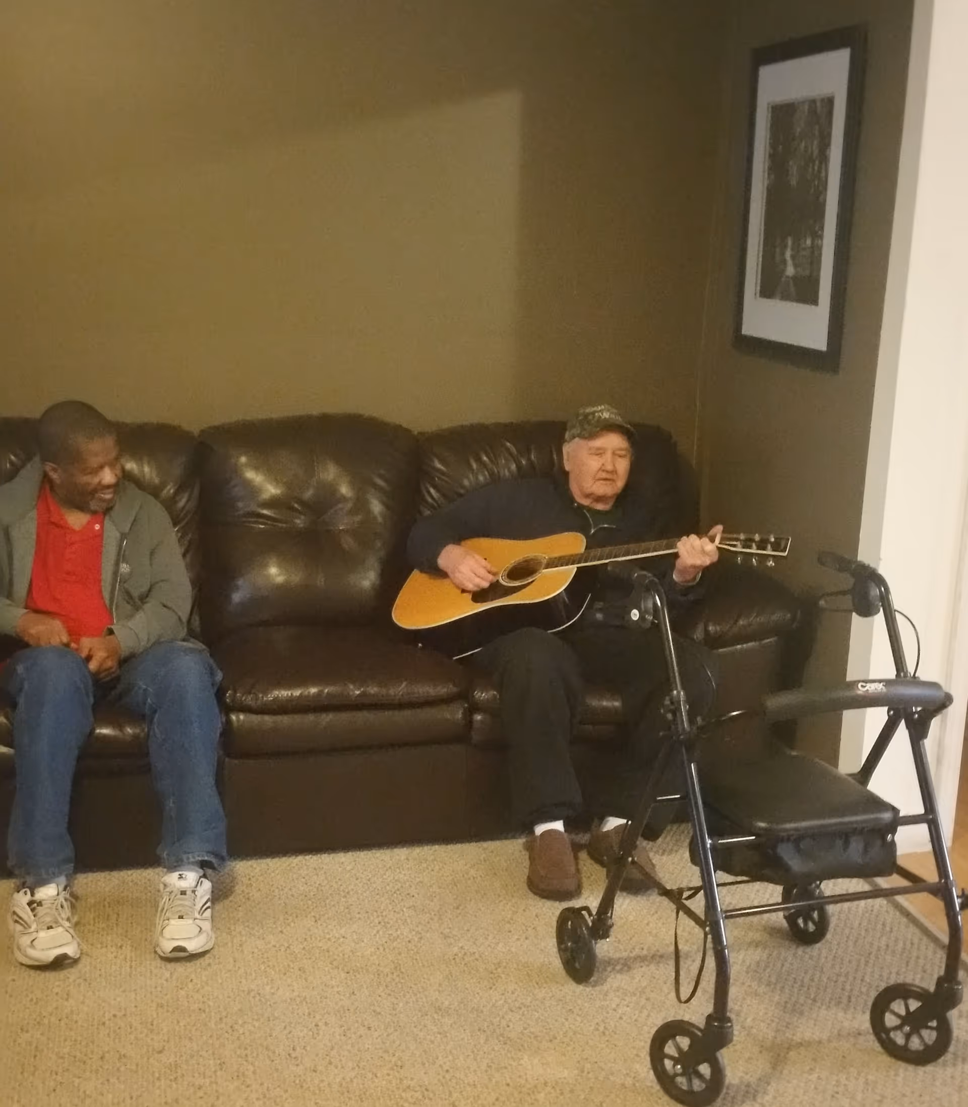 Two men sitting on a dark brown leather couch in a living room. One man is playing an acoustic guitar while the other man watches and smiles. A black walker is positioned in front of the man playing the guitar. There is a framed black and white picture hanging on the wall behind them.