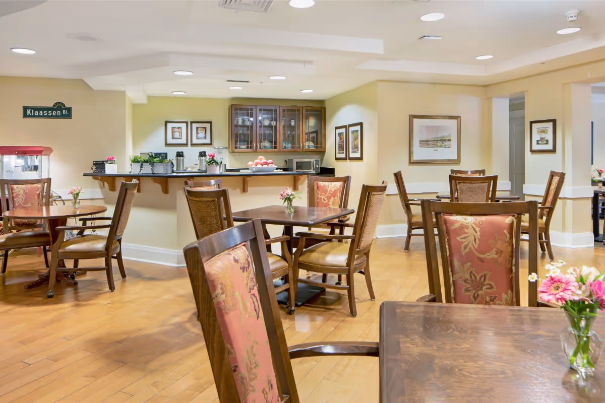 Bright dining room with multiple wooden tables and upholstered chairs arranged around a central serving counter and small floral centerpieces.