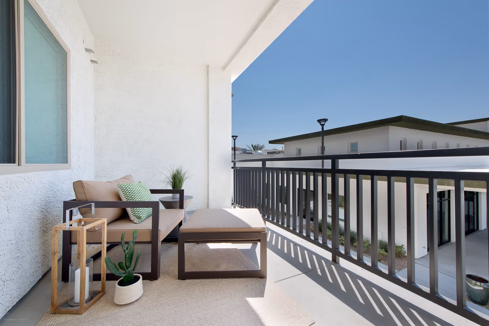 A bright outdoor balcony area with a cushioned armchair and matching ottoman, a green patterned pillow, a small potted cactus, a wooden lantern with a candle, and a small plant on a side table. The balcony has a black metal railing and overlooks a modern building under a clear blue sky.