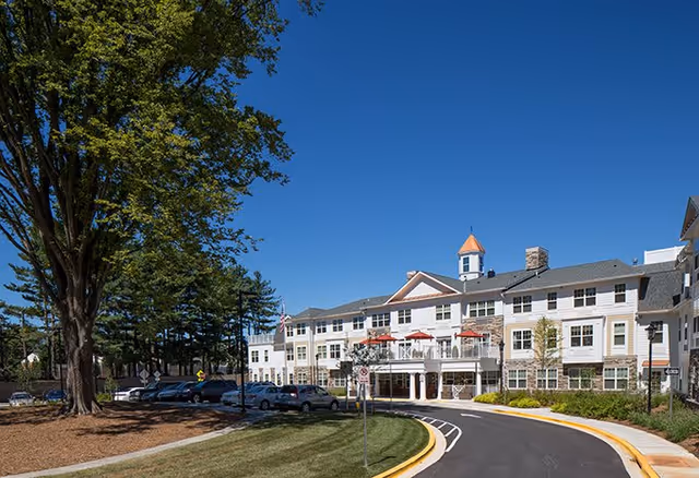 Exterior view of Brightview Fallsgrove senior assisted living and memory care facility with a clear blue sky, a curved driveway leading to the entrance, parked cars, and large trees on the left side.