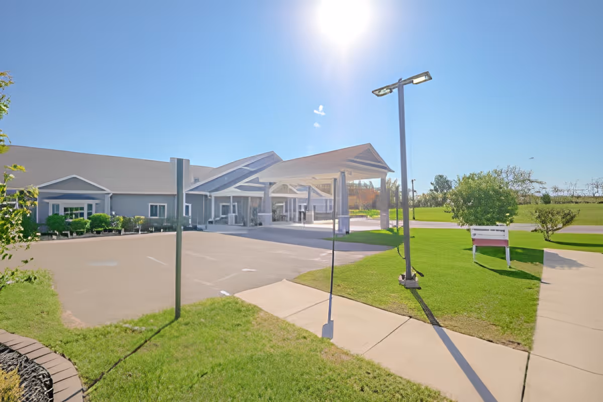 Exterior view of Quality Care of Howell facility on a sunny day, showing a paved parking area, green lawn with a bench, a tall street lamp, and the building entrance with a covered drop-off area.