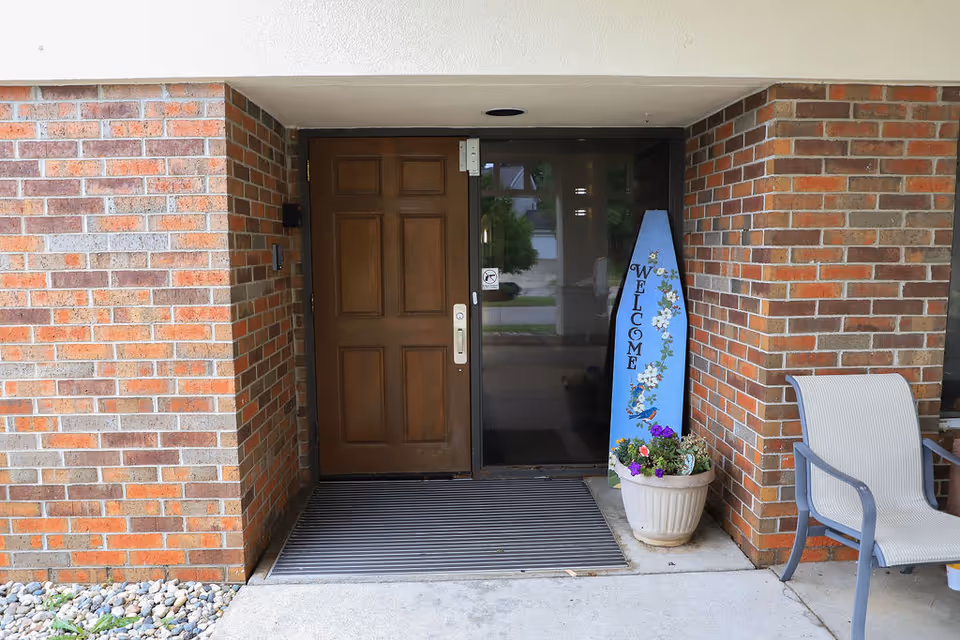 Entrance to a building with a brown wooden door and a glass panel beside it. A blue vertical welcome sign decorated with flowers and a bird is placed next to a white planter with colorful flowers. There is a beige outdoor chair to the right, and the walls are made of red and brown bricks.