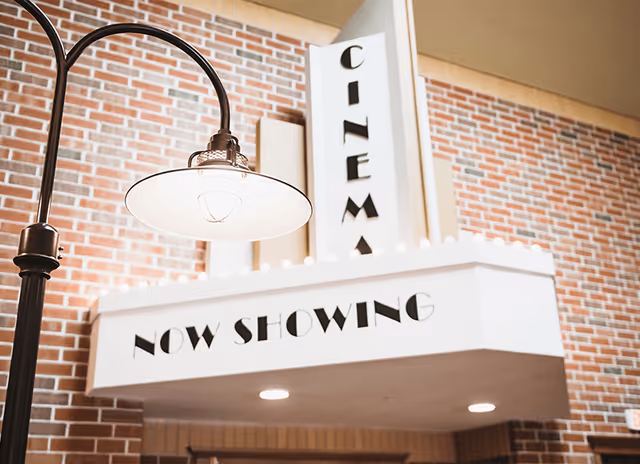 Interior view of a cinema marquee sign with the words 'CINEMA' and 'NOW SHOWING' displayed, mounted on a brick wall with a vintage-style lamp hanging nearby.