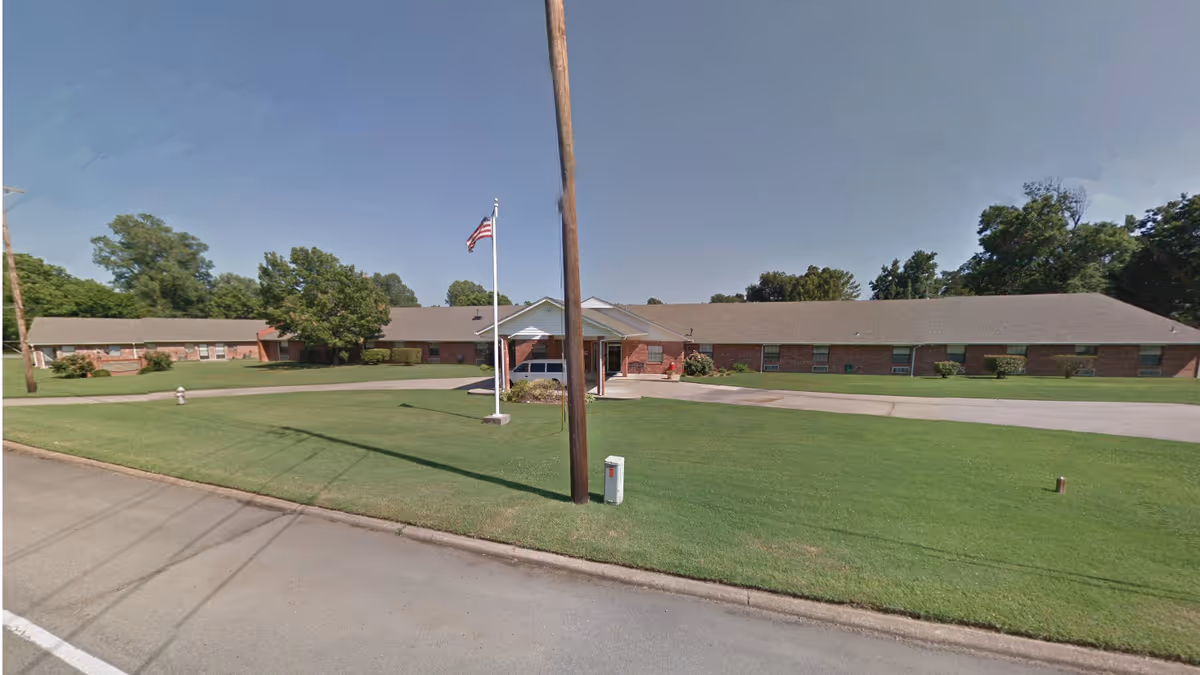 Low one-story brick nursing home building with a covered entrance, flagpole, driveway and well-kept lawn.