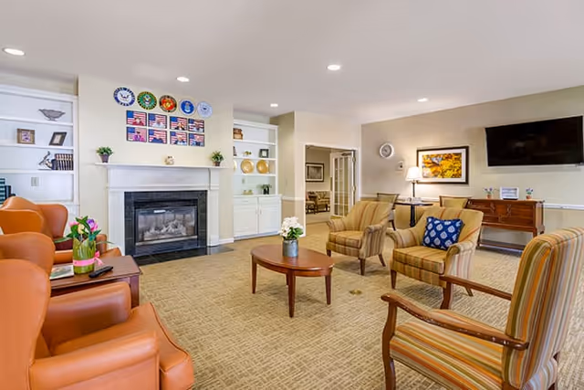 A cozy living room area with a fireplace decorated with military insignias and American flags above it. The room has several comfortable chairs arranged around a wooden coffee table with a small plant on it. There are built-in shelves with decorative items, a wall-mounted TV, a side table with a lamp, and framed artwork on the walls. The carpet is beige, and the room is well-lit with recessed ceiling lights.