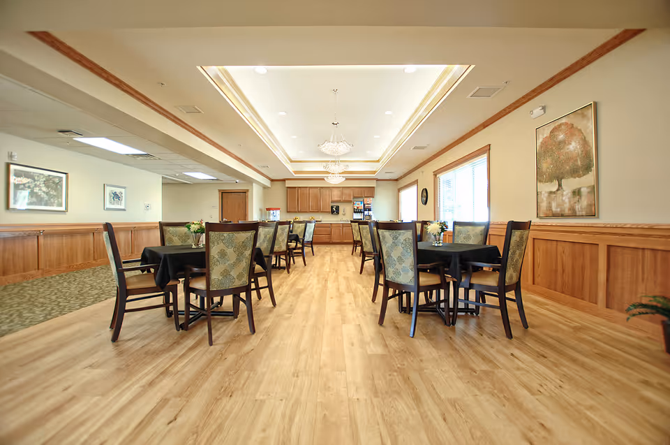 Bright communal dining room with multiple round tables and upholstered chairs in a senior living facility.