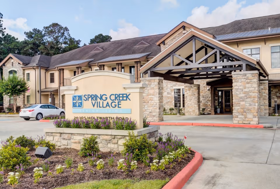 Exterior view of Spring Creek Village senior living facility showing the main entrance with a covered driveway, stone pillars, and a sign with the facility name and address surrounded by landscaped flowers and greenery.