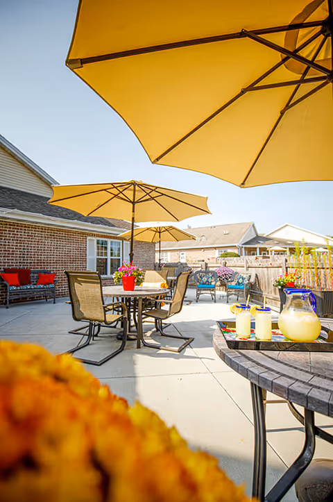 Outdoor patio area at Frontida Assisted Living: Clifden Court with tables, chairs, large yellow umbrellas, and a pitcher of lemonade with glasses on a table. There are flowers in the foreground and seating areas with cushions near a brick building under a clear blue sky.