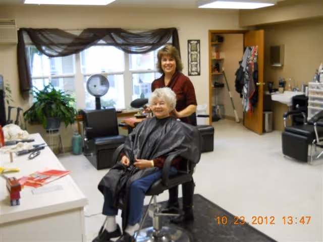 An elderly woman is sitting in a salon chair wearing a black haircut cape, smiling while a hairstylist stands behind her in a well-lit room with salon equipment and chairs.