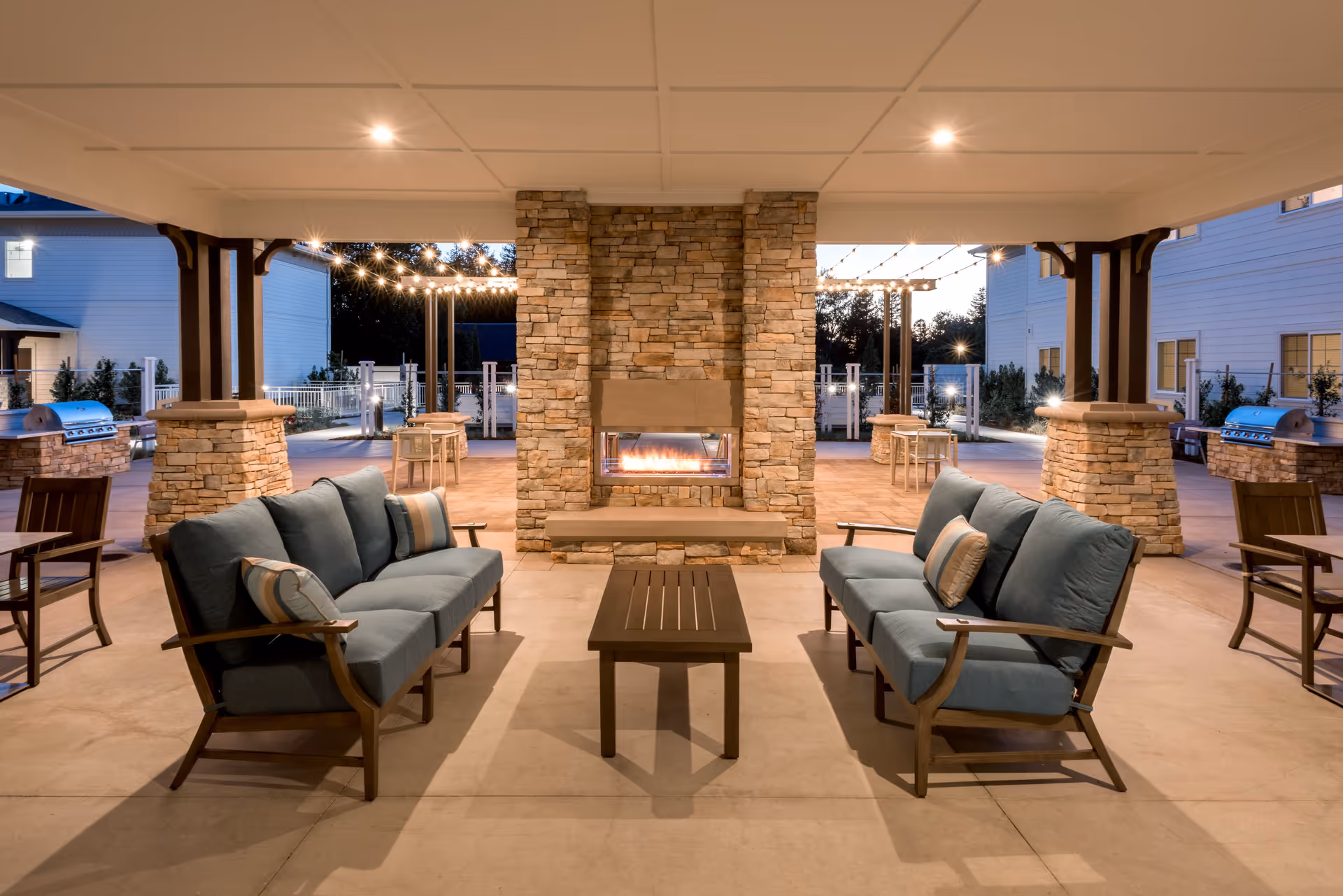 Outdoor covered seating area with two cushioned sofas facing each other and a rectangular coffee table in between, centered around a stone fireplace. String lights hang above, and there are stone pillars and barbecue grills in the background with residential buildings visible.