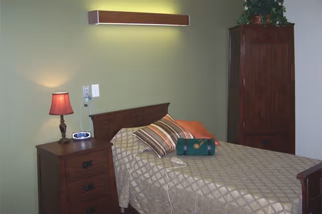 A bedroom with a single bed covered with a beige patterned bedspread and several pillows, including striped and solid-colored ones. Next to the bed is a wooden nightstand with a red lampshade and a small clock. A tall wooden cabinet with a plant on top is positioned against the wall. The walls are painted light green and white.
