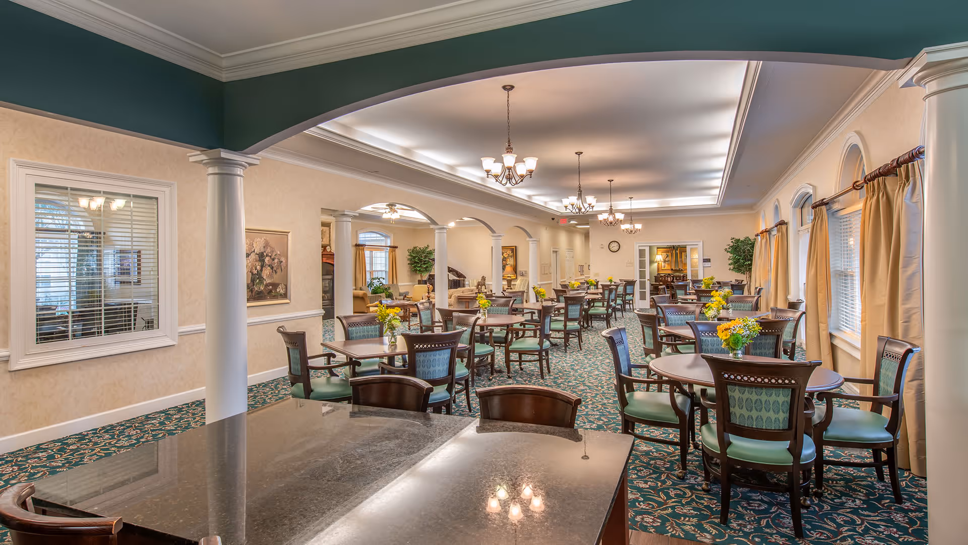 A spacious dining room with multiple round tables and chairs arranged neatly. Each table has a small vase with yellow flowers. The room features green patterned carpet, cream-colored walls, large windows with beige curtains, white columns, and elegant chandeliers hanging from the ceiling. There is a seating area with sofas and plants visible in the background.
