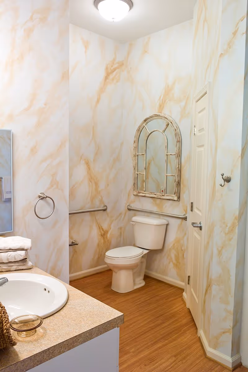A bathroom with beige and light brown marbled walls, a white toilet with grab bars on both sides, a rustic arched mirror above the toilet, a white sink with a beige countertop, folded towels, and a towel ring on the wall.