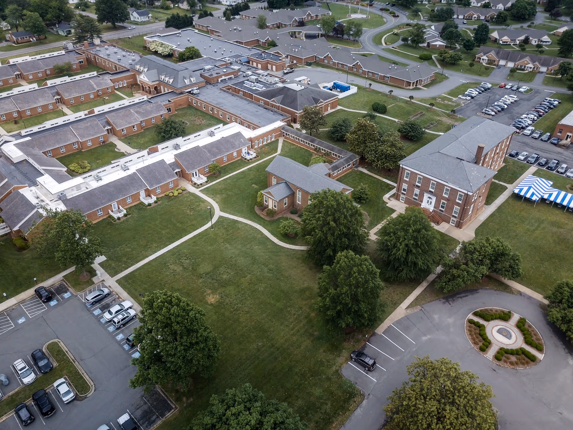 Aerial view of the Masonic Home of Virginia facility showing multiple brick buildings with gray roofs, green lawns, trees, walkways, parking lots with cars, and a blue and white striped tent on the grass.