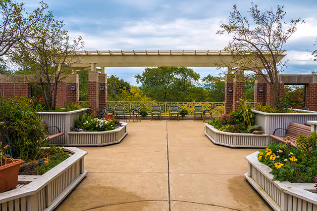 Outdoor patio area with built-in planter benches containing flowers and small trees, brick pillars supporting a pergola structure, and several chairs arranged along a railing overlooking greenery under a cloudy sky.