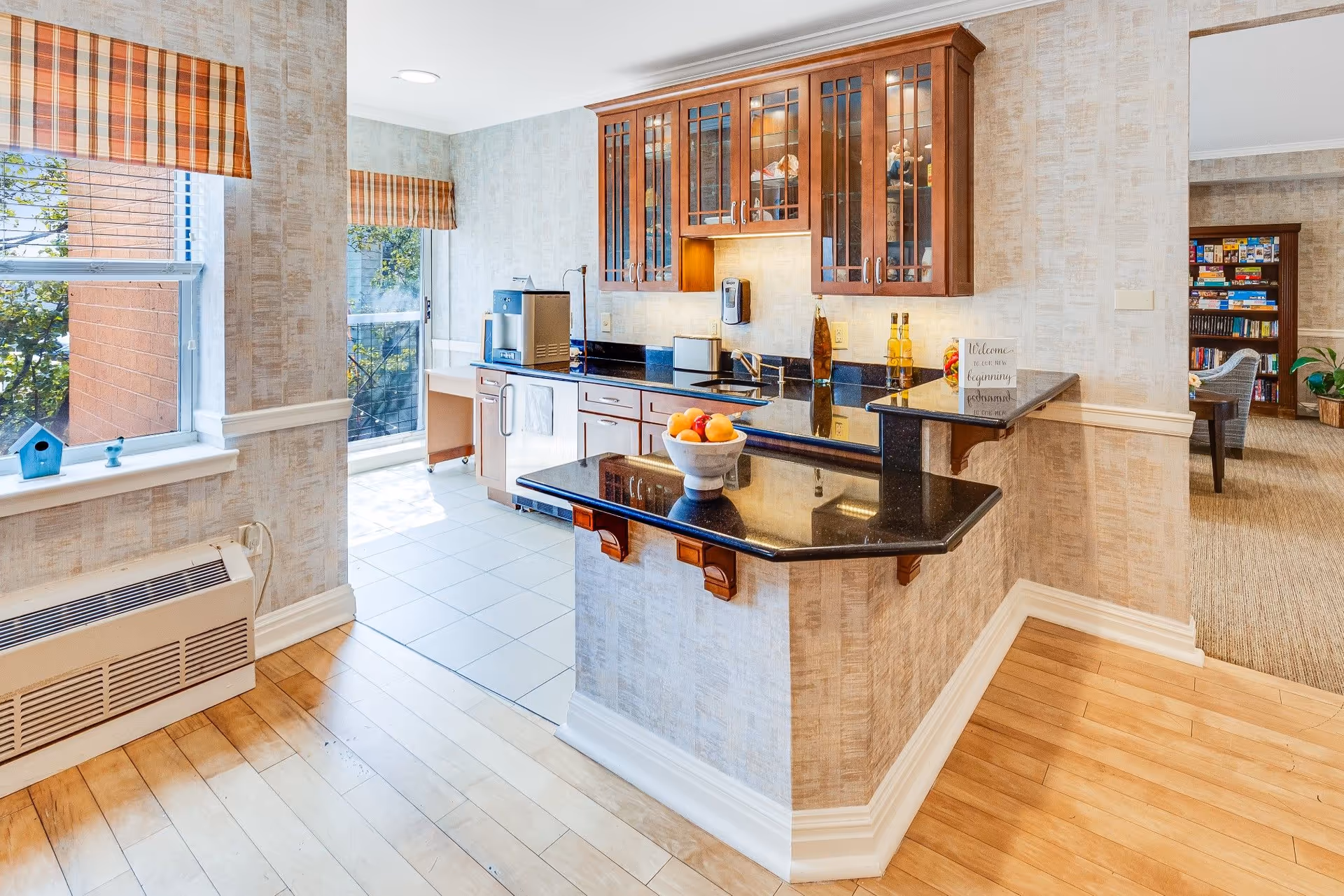Bright kitchen area with wooden cabinets featuring glass doors, black granite countertops, a sink, and a small breakfast bar with a bowl of fruit. The kitchen has tiled flooring and a window with striped valances letting in natural light. Adjacent to the kitchen is a carpeted room with a bookshelf filled with board games and a wicker chair.