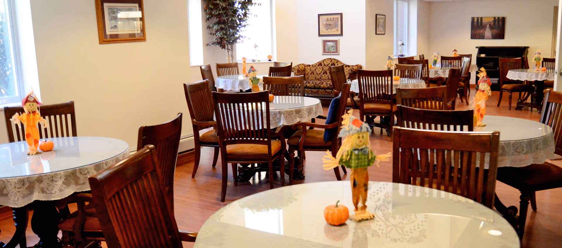 Bright dining room with multiple round tables and wooden chairs, each table decorated with small scarecrow and pumpkin centerpieces.
