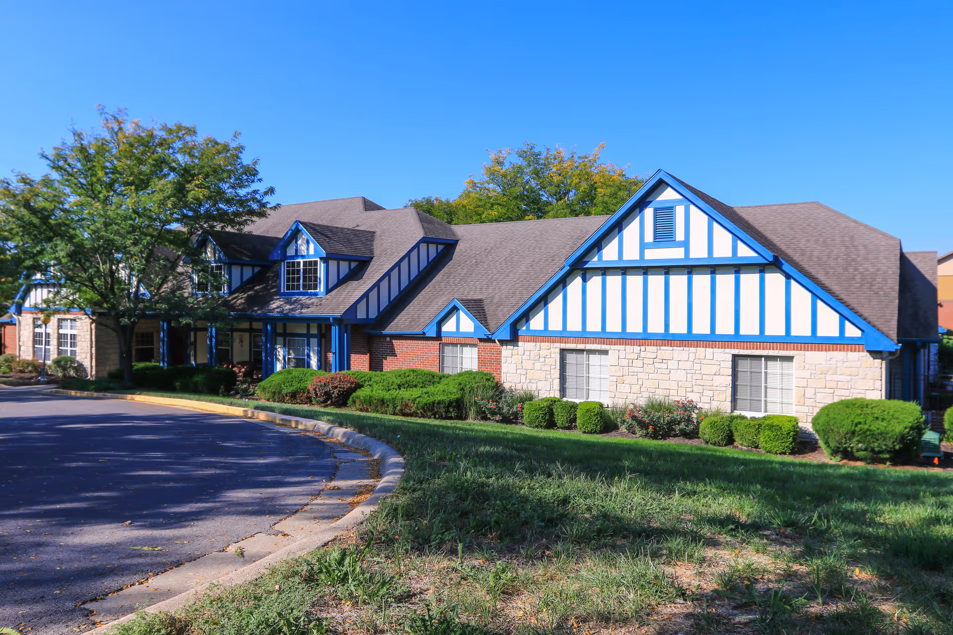 Exterior view of a senior living facility building with Tudor-style architecture featuring blue and white trim, brick and stone walls, surrounded by green bushes and trees under a clear blue sky.