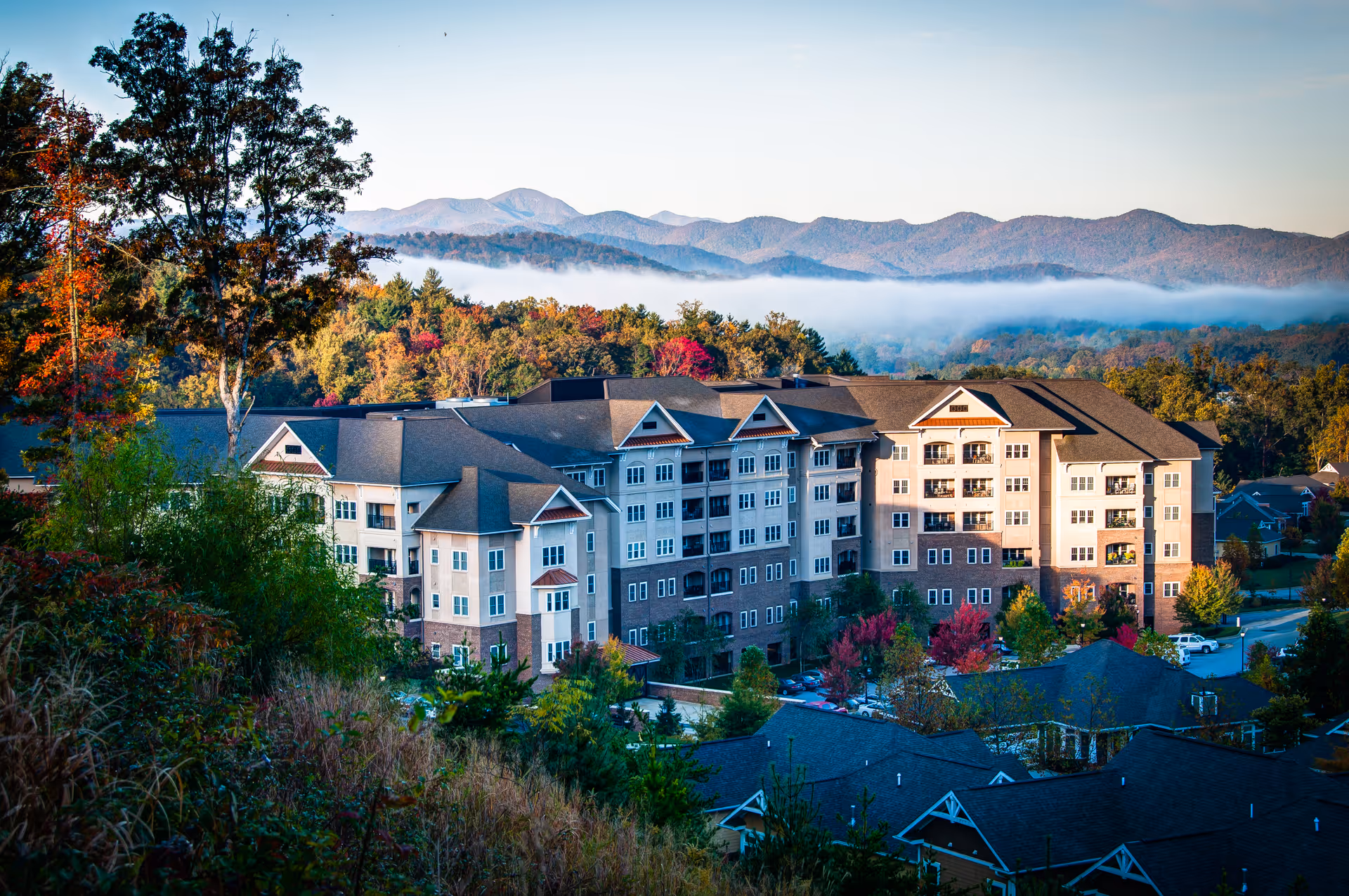 A large multi-story senior living building surrounded by trees with mountains and morning fog in the background.