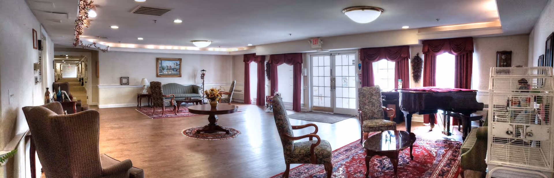 A spacious assisted living common area with wooden flooring, multiple seating arrangements including armchairs and sofas, a round wooden table with a floral centerpiece, a grand piano near large windows with red curtains, and a birdcage on the right side. The room is well-lit with ceiling lights and natural light from the windows.