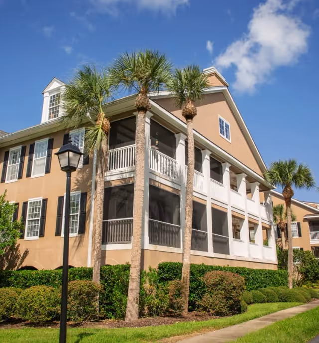Exterior view of a two-story beige building with white columns and screened balconies, surrounded by palm trees and well-maintained bushes under a blue sky with some clouds.