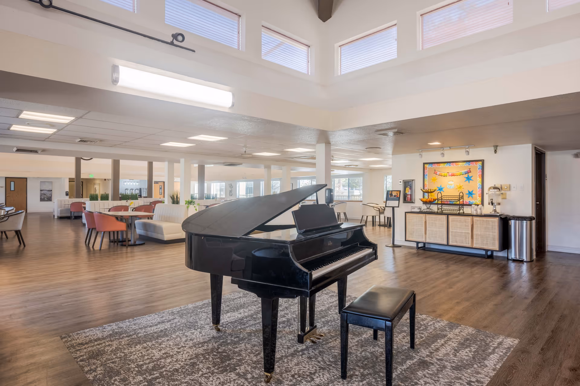 Spacious senior living facility common area with a black grand piano and matching bench on a patterned rug in the foreground. The room features wood flooring, multiple seating areas with chairs and sofas, large windows, and a bulletin board on the wall. The ceiling has high windows allowing natural light to enter.