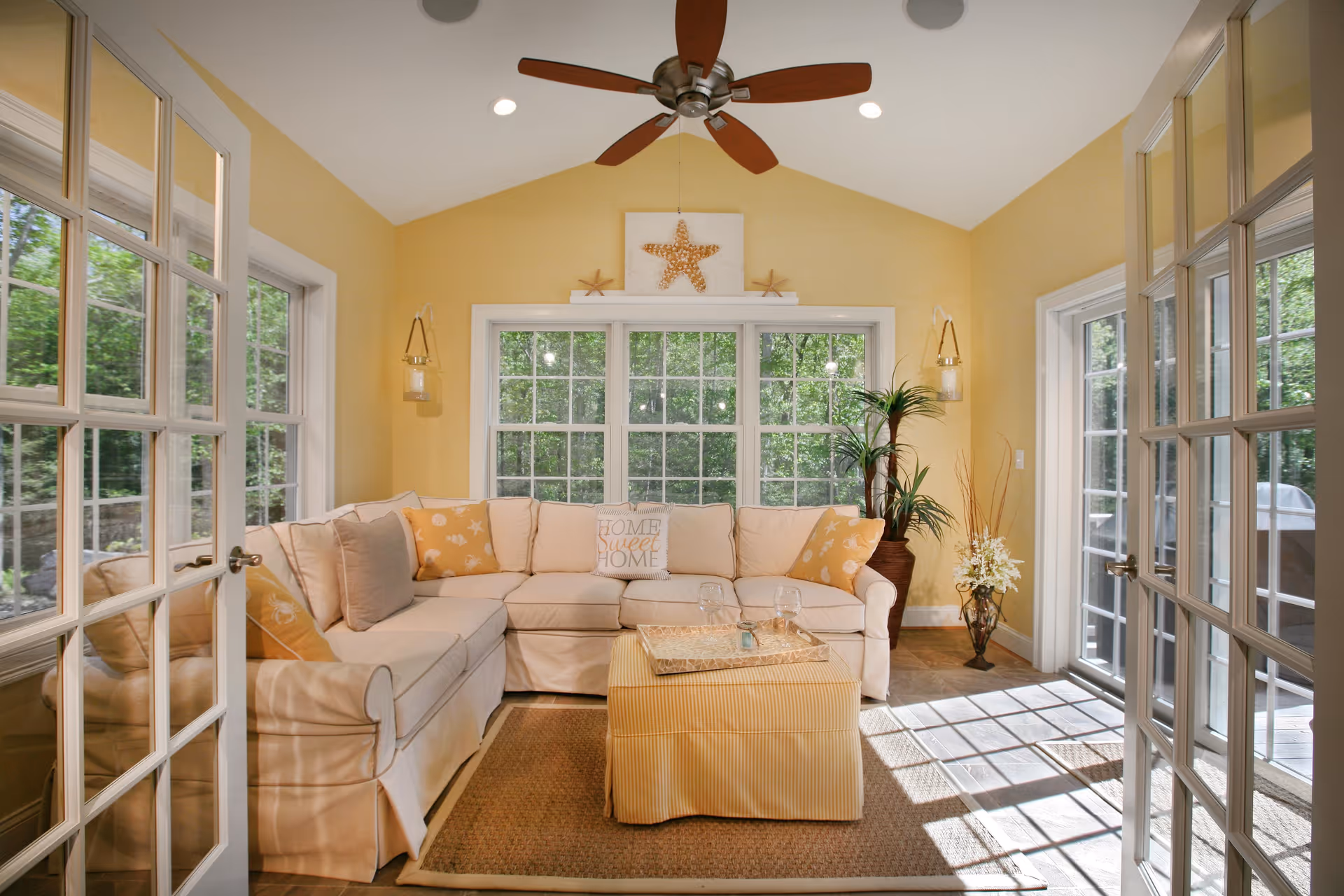 Bright sunlit living room with a beige sectional sofa, ottoman coffee table, large windows and French doors.