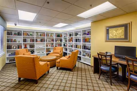 Bright library lounge in a senior facility with orange chairs, white bookshelves, and a computer desk.