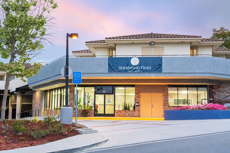 Exterior view of Sherwood Oaks Post Acute facility at dusk, showing the entrance with large windows, a blue banner with the facility name, a street lamp, landscaped plants, and a clear sky with pink and purple hues.