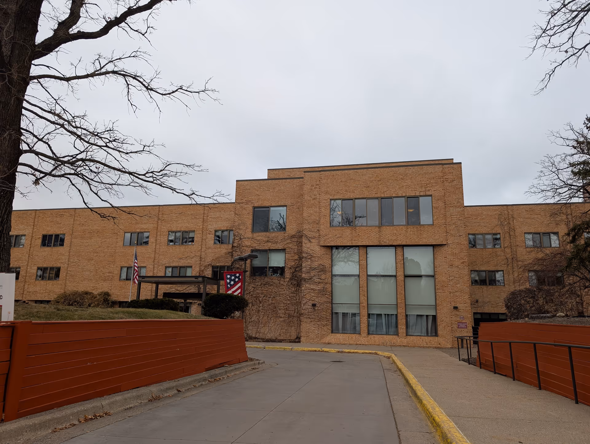 Three-story brick senior living building with large windows, an entrance canopy, driveway, and American flags.