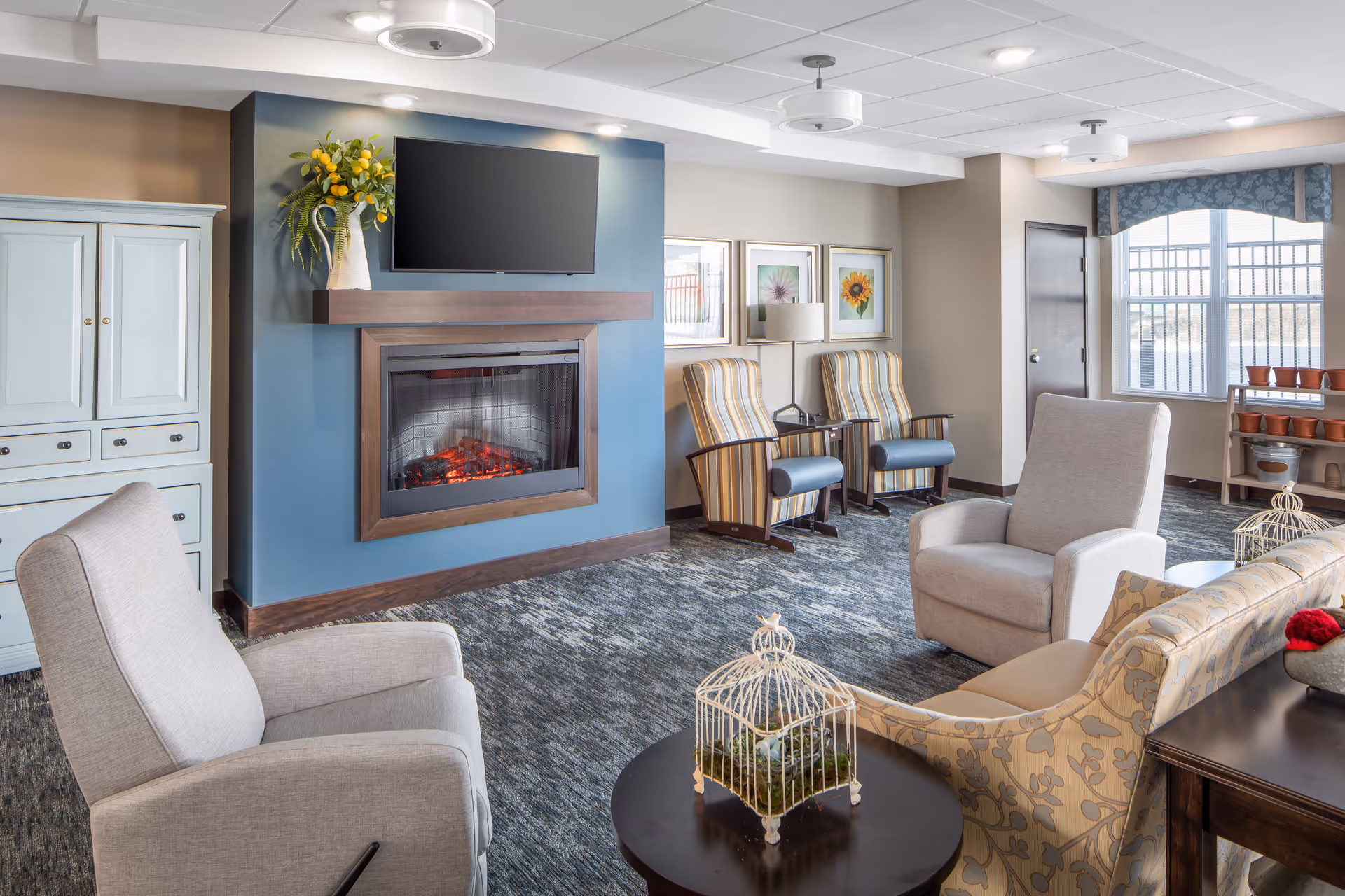 A cozy living room area in a senior living facility featuring a modern electric fireplace with a TV mounted above it. The room has comfortable armchairs and a patterned sofa arranged around a round coffee table with decorative birdcages. There are two striped armchairs with a small table and lamp between them near the wall, which is adorned with framed floral artwork. A window with a blue valance lets in natural light, and a white cabinet stands against one wall.