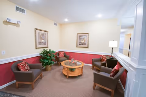 A cozy seating area with four brown armchairs arranged around a round wooden coffee table. The chairs have red patterned cushions. The walls are painted yellow with a red wainscoting and white trim. Two framed pictures hang on the wall, and a potted plant is placed in the corner. A table lamp is on a small side table next to one of the chairs.