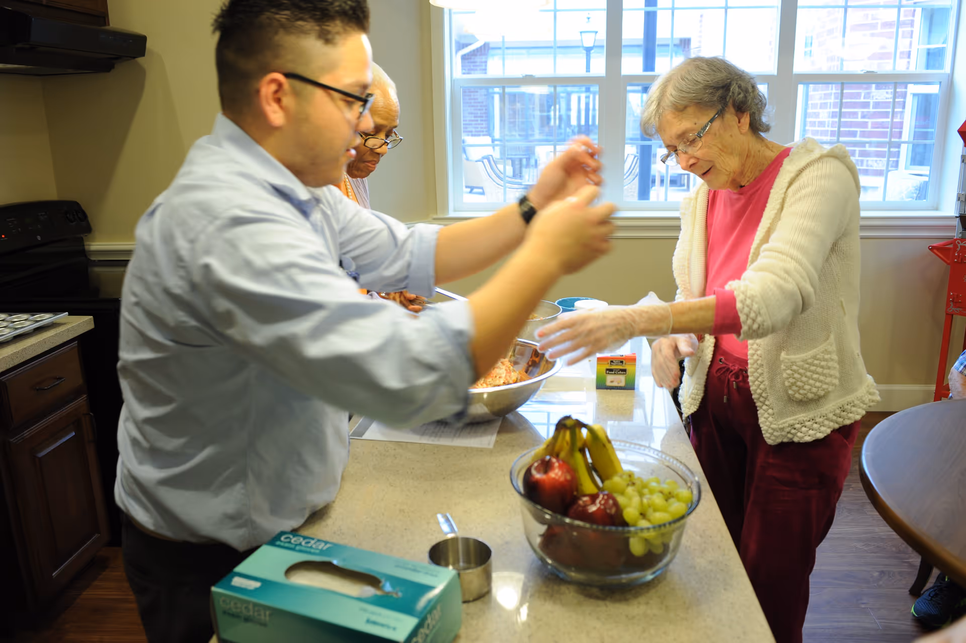 A man and two elderly women are preparing food together in a kitchen. The man is in the foreground, wearing glasses and a light blue shirt, while one elderly woman in a white cardigan and pink shirt is reaching towards the counter. A bowl of fruit including bananas, apples, and grapes is on the counter, along with a box of gloves and a measuring cup. The kitchen has wooden cabinets and a window showing an outdoor patio area.