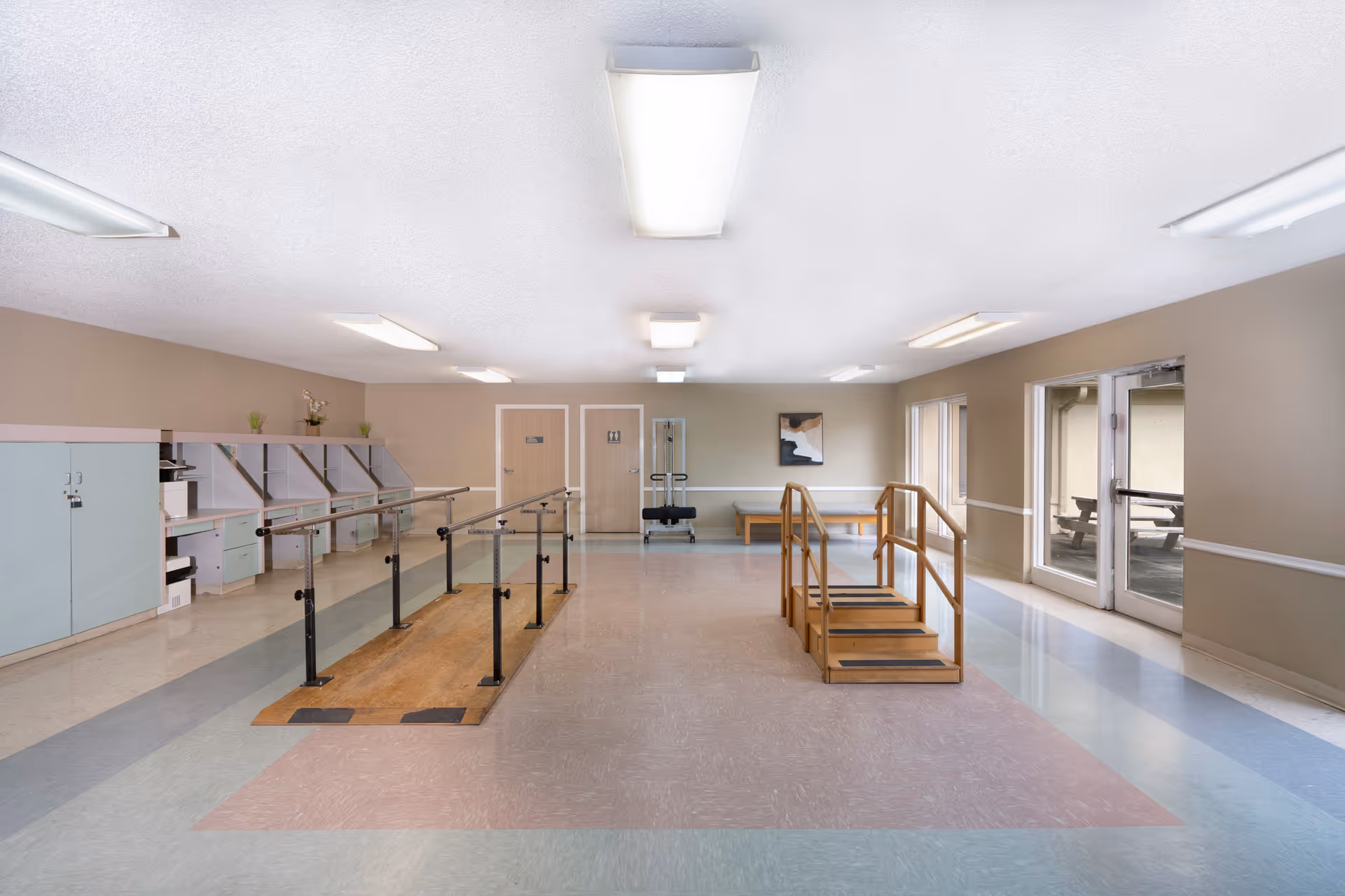 Spacious interior rehabilitation room with parallel walking bars and wooden practice stairs, cabinets and seating along the walls.