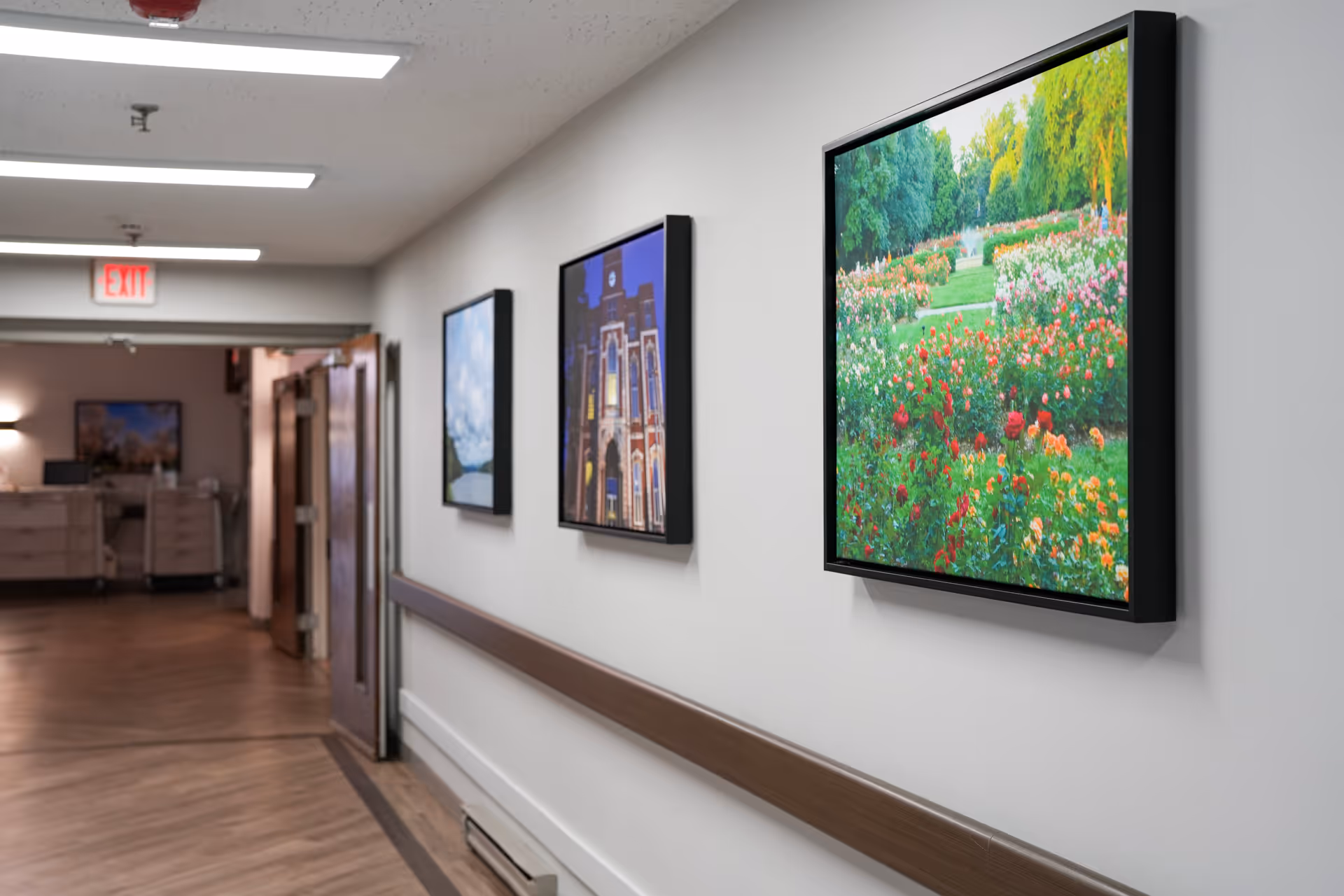 A hallway in a senior living facility with framed pictures on the wall, including a colorful garden scene and a building. The floor is wooden, and there is a handrail along the wall. At the end of the hallway, there is an exit sign above a door and some furniture visible in the background.