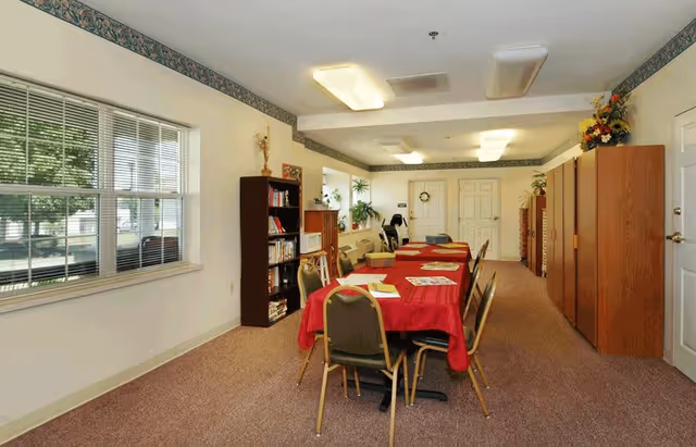 A long room with a table covered in a red tablecloth surrounded by chairs. The room has beige carpet, white walls with a floral border near the ceiling, and several wooden cabinets and bookshelves along the walls. Large windows on the left side let in natural light, and there are ceiling lights providing additional illumination.