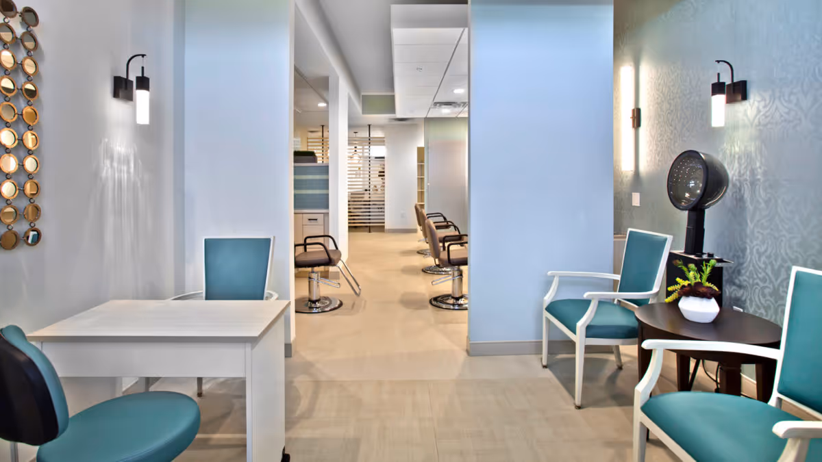 Interior salon area with teal-upholstered chairs, a manicure table, and a hooded hair dryer beside a row of styling stations.