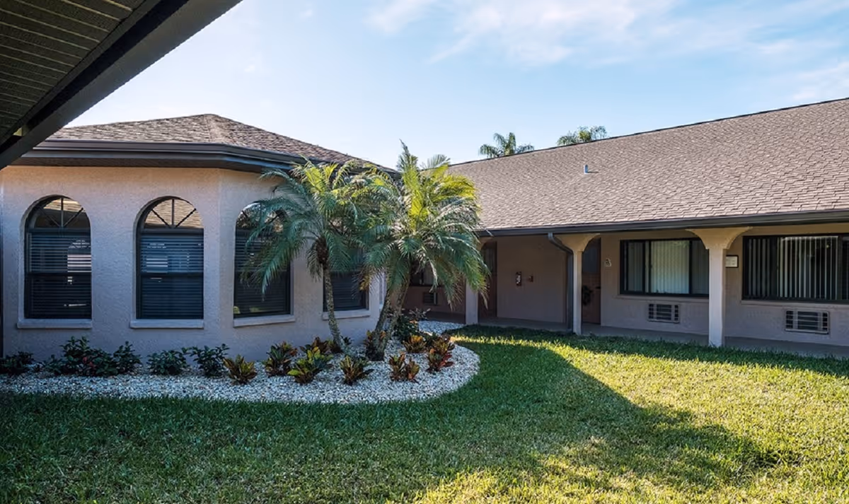 Outdoor courtyard area of a senior living facility with a well-maintained lawn, palm trees, and shrubs surrounded by a building with arched windows and covered walkways under a clear blue sky.